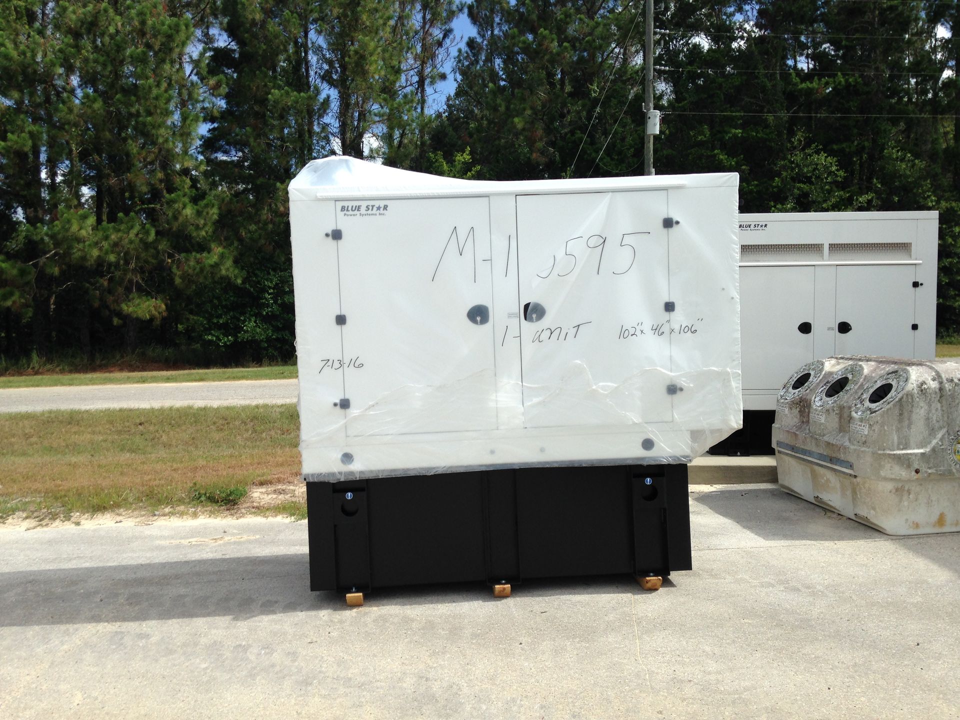 White industrial generator or equipment on a black base outdoors beside trees and other machinery.
