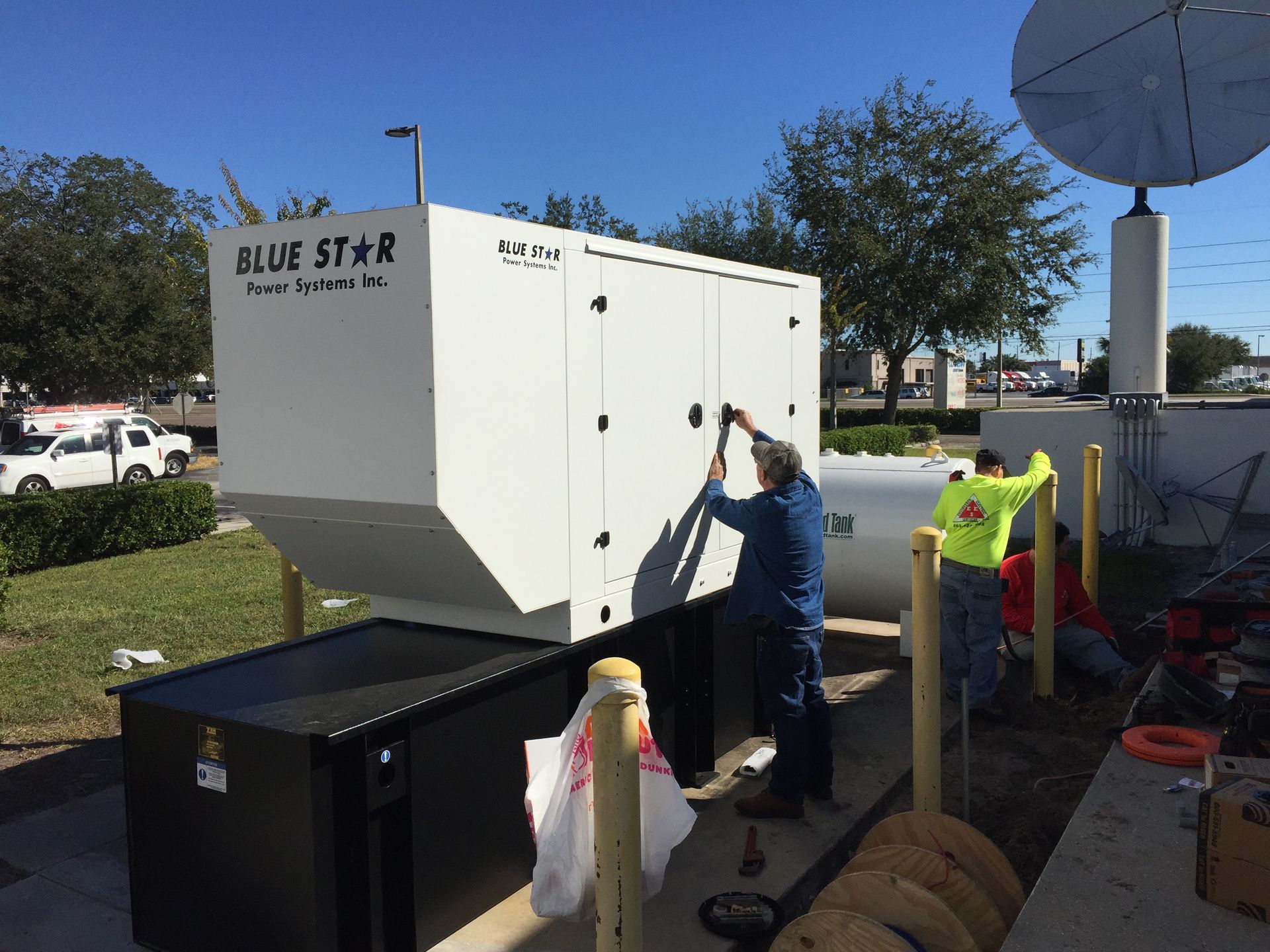 Worker refuels a white generator at a roadside work site, with cones and equipment nearby.