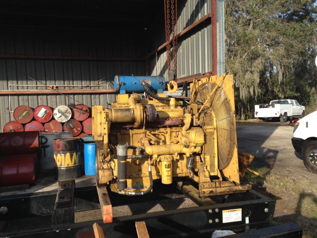 Yellow industrial engine on a trailer beside a barn, with blue pipework and red barrels in the background.