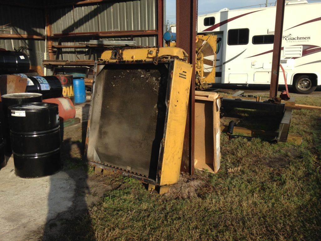 Yellow backhoe bucket and black barrels outside a corrugated metal shed, with a white RV in the background