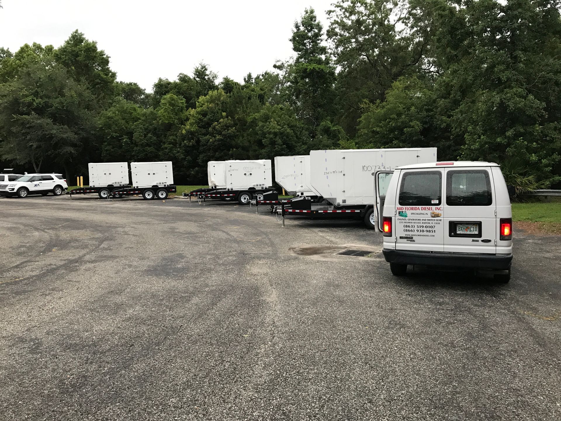 White vans and trailers parked on a gravel lot beside trees under an overcast sky.