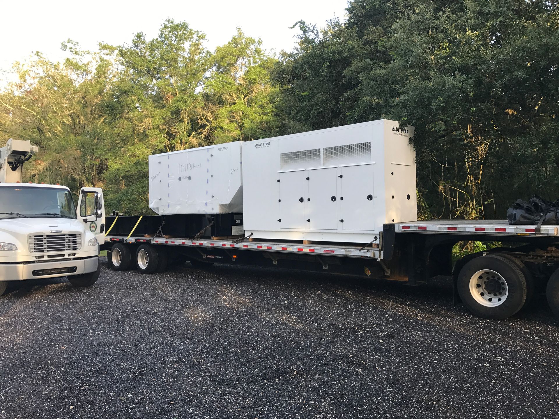 White box truck parked beside a flatbed trailer on a gravel lot, with trees in the background