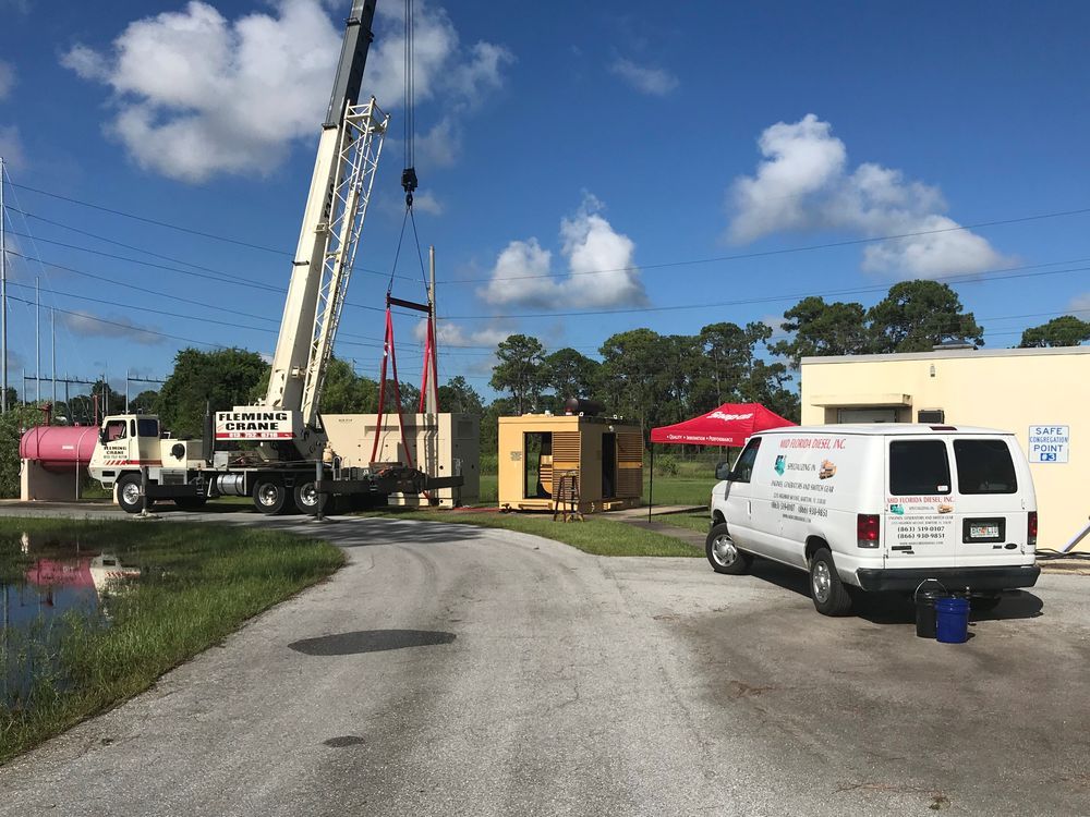 Crane truck lifting a tall pole at a work site, with vans and equipment beside a gravel road under a blue sky.