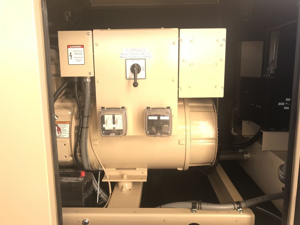 Beige industrial generator inside an enclosed utility compartment, with pipes and wiring visible.