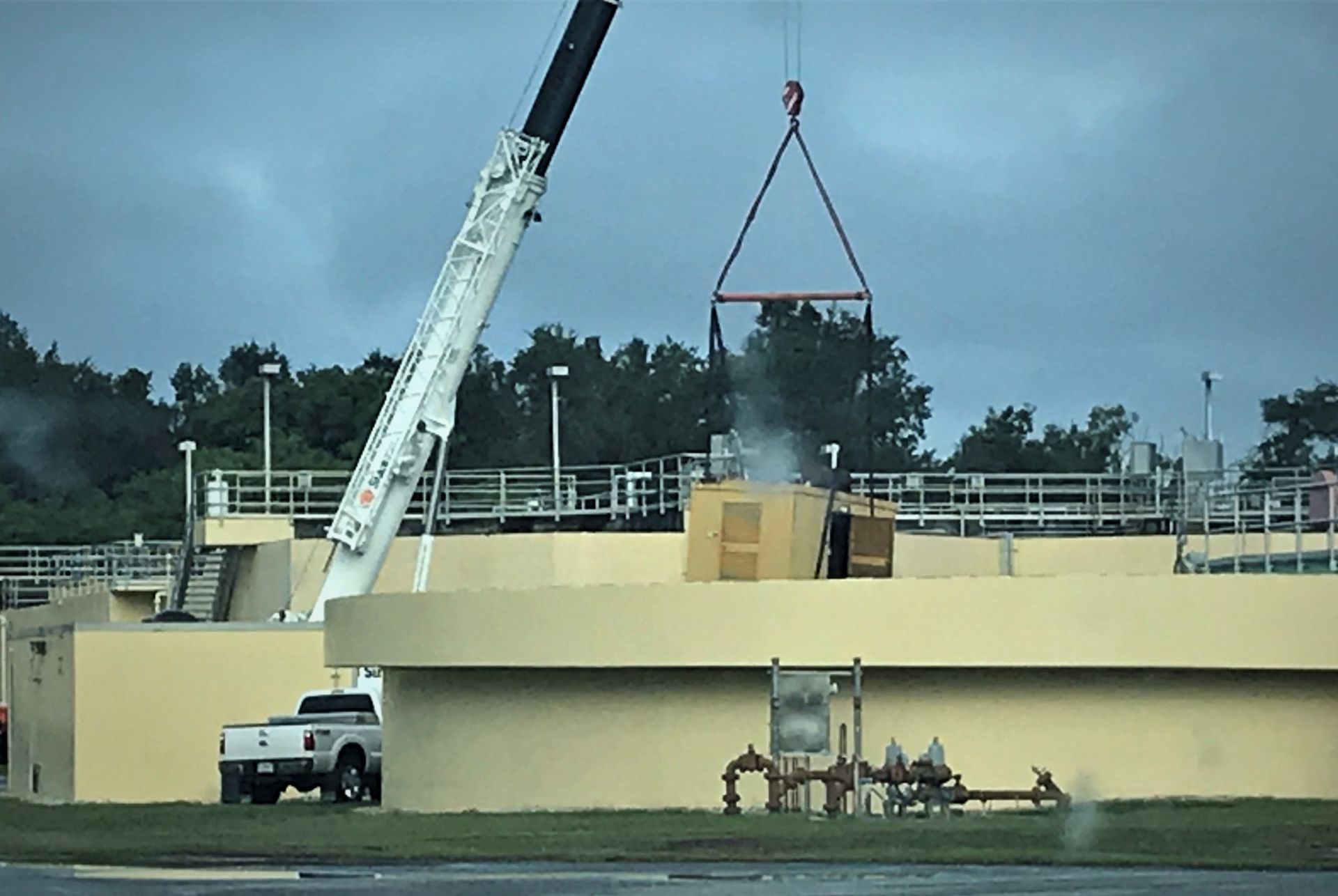 Crane hoisting a large object over a beige industrial building beside a parked white truck