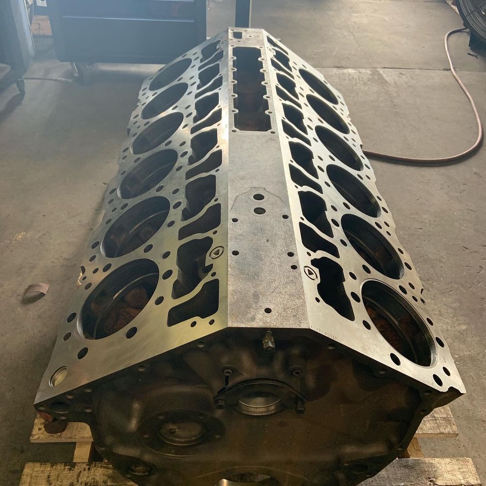 Large metal engine block on a workshop floor, viewed from above, with cylinder bores exposed.