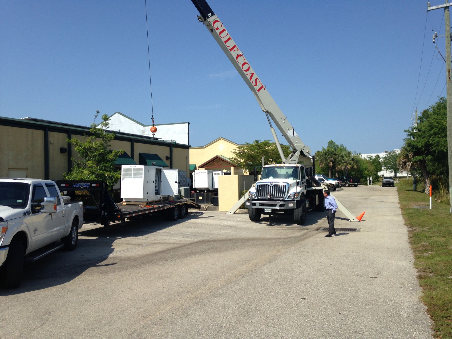 Crane truck lifting a white cabinet beside a building on a sunny gravel lot