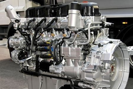 Silver automotive engine displayed on a stand in a workshop or factory setting