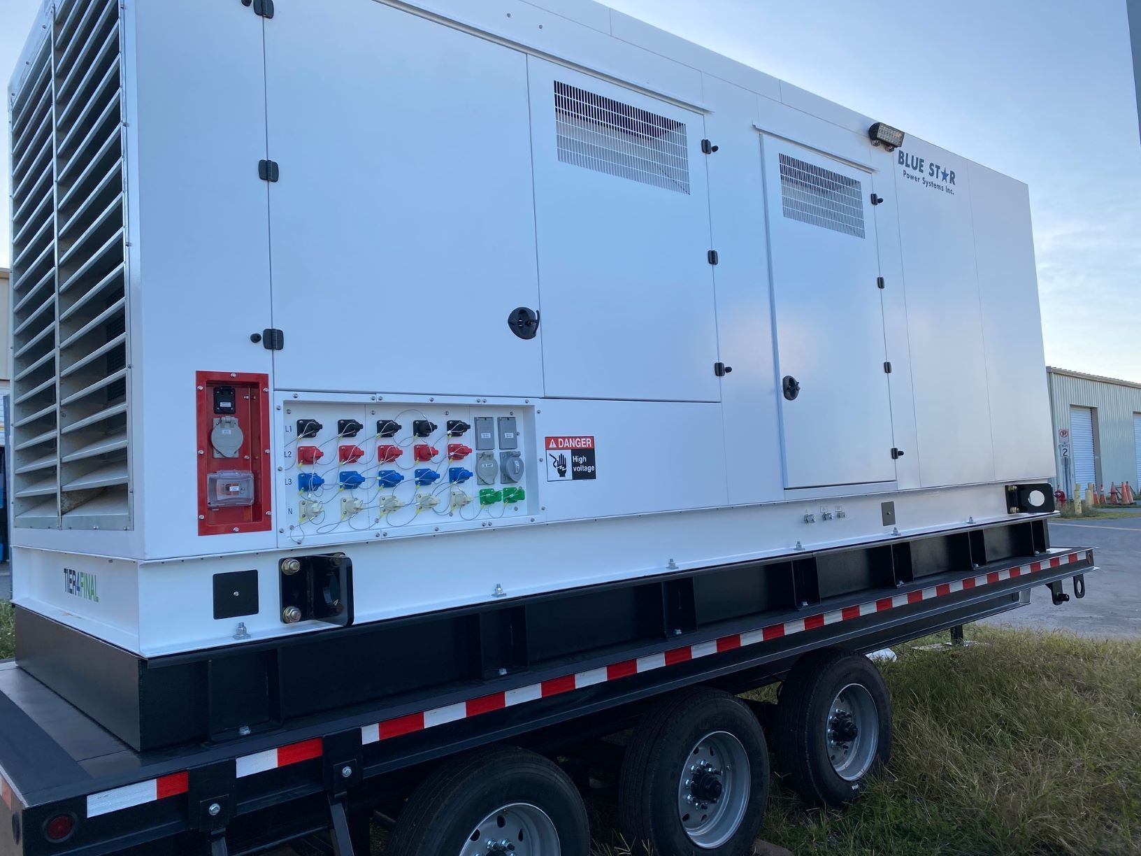 White industrial generator on a black trailer in a grassy field, with control panel and vents visible
