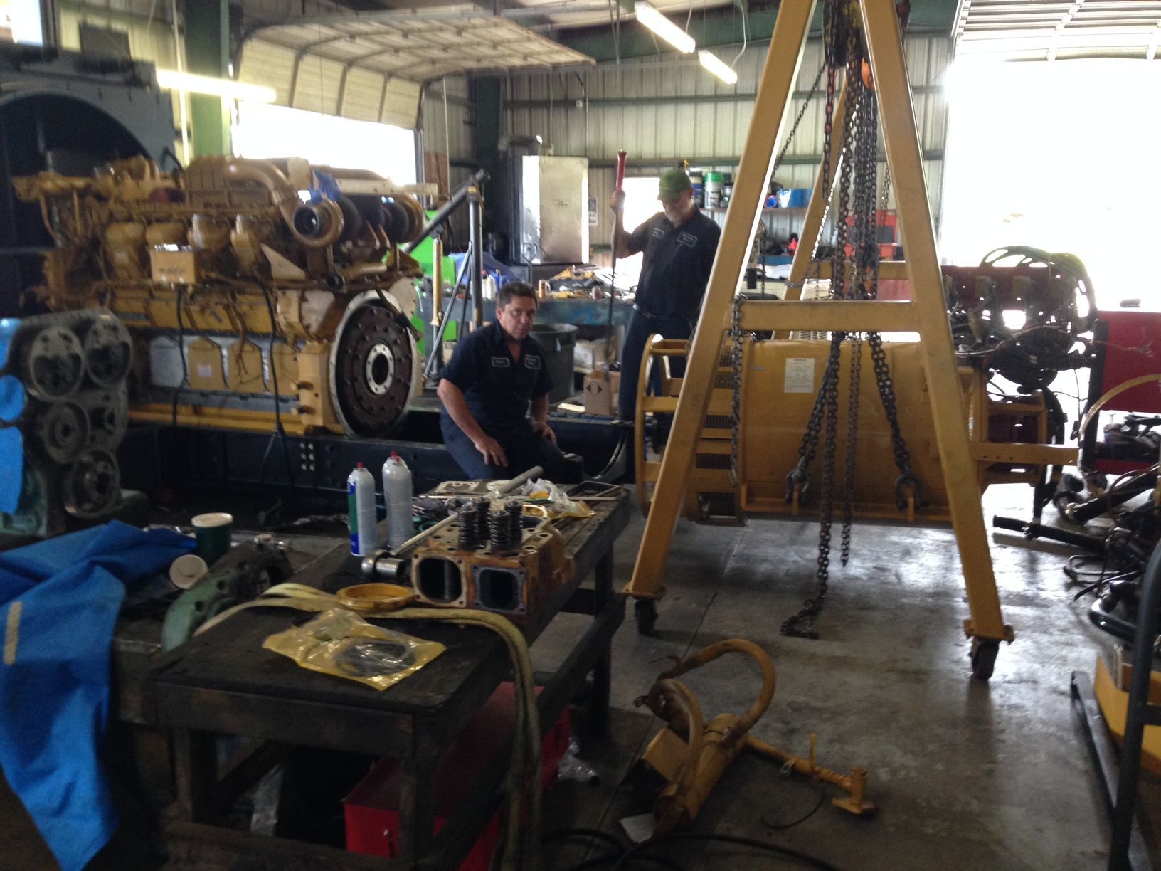 Mechanic working in a cluttered auto repair shop beside a lifted engine block and tools