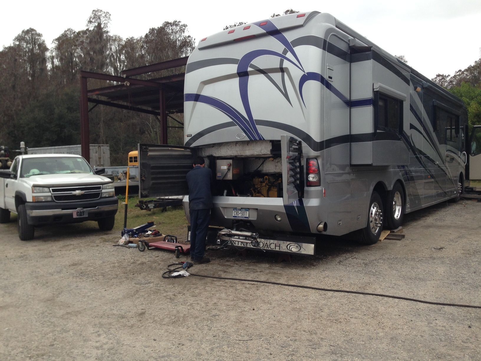 Person working on the rear of a silver RV in a lot, with tools and a pickup truck nearby