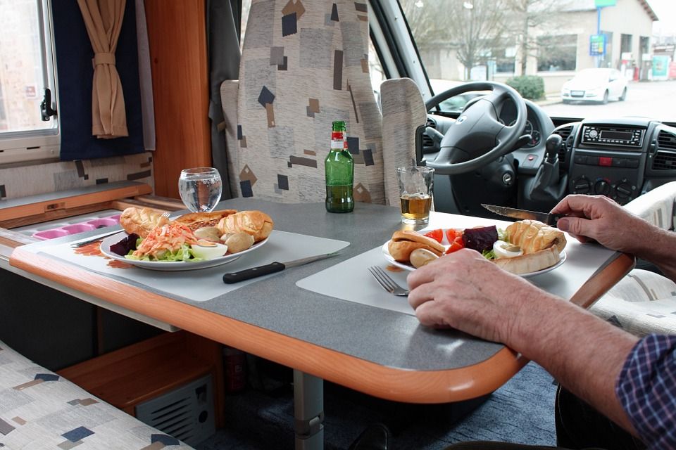 Two people eating lunch at a camper van table, with sandwiches, drinks, and a steering wheel visible.
