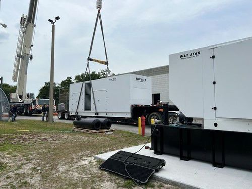 Utility equipment being hoisted by crane at an outdoor work site with trucks and trailers nearby