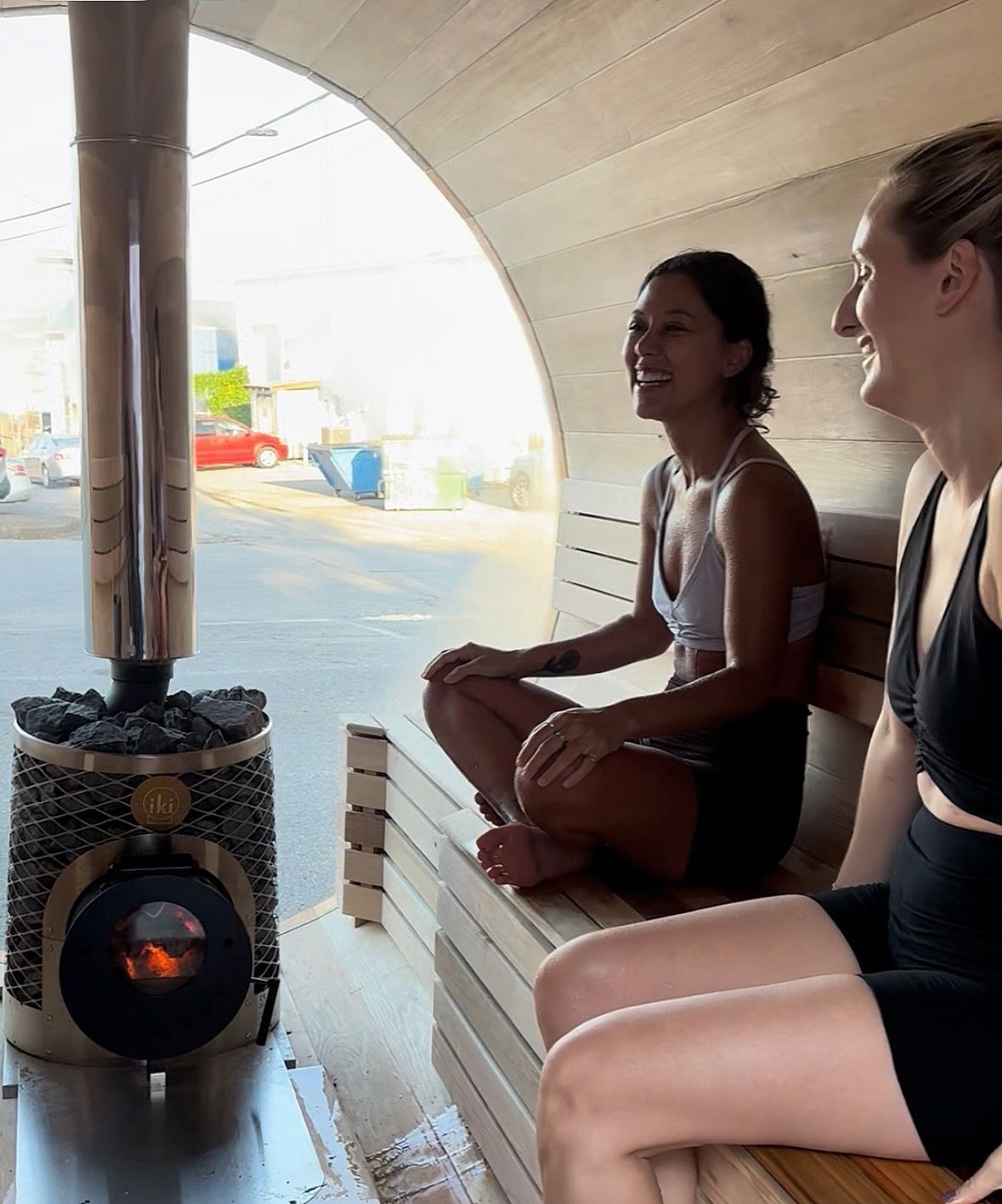Two people laughing in a wooden sauna, near a lit stove. Bright outdoors visible through opening.