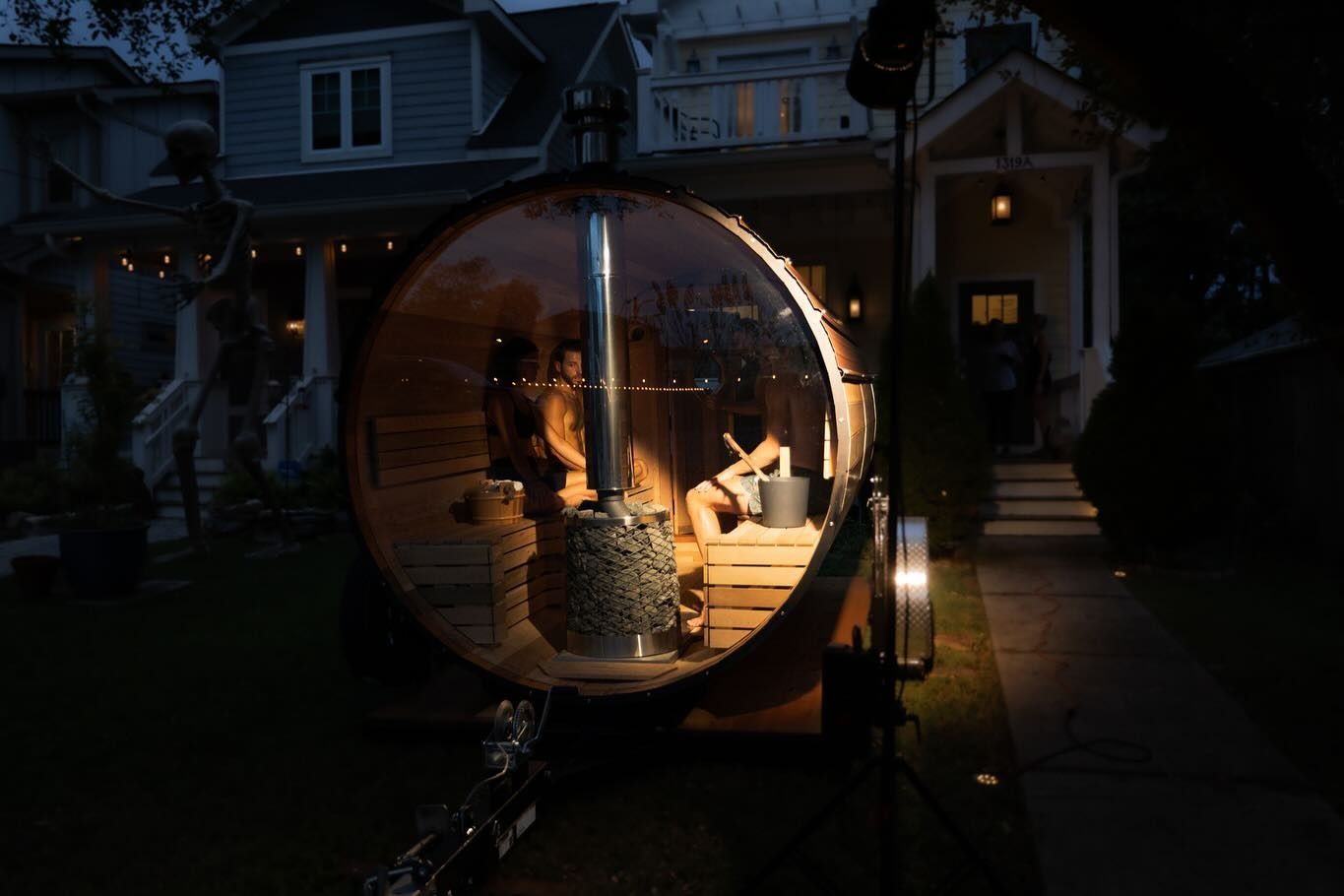 A barrel sauna with people inside, lit up at night in a yard with houses in the background.