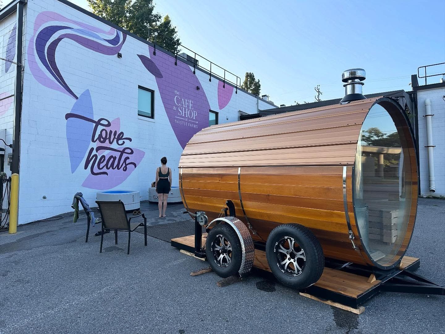 Wooden barrel sauna on wheels beside a building with a mural that says "love heals." A person walks towards the sauna.