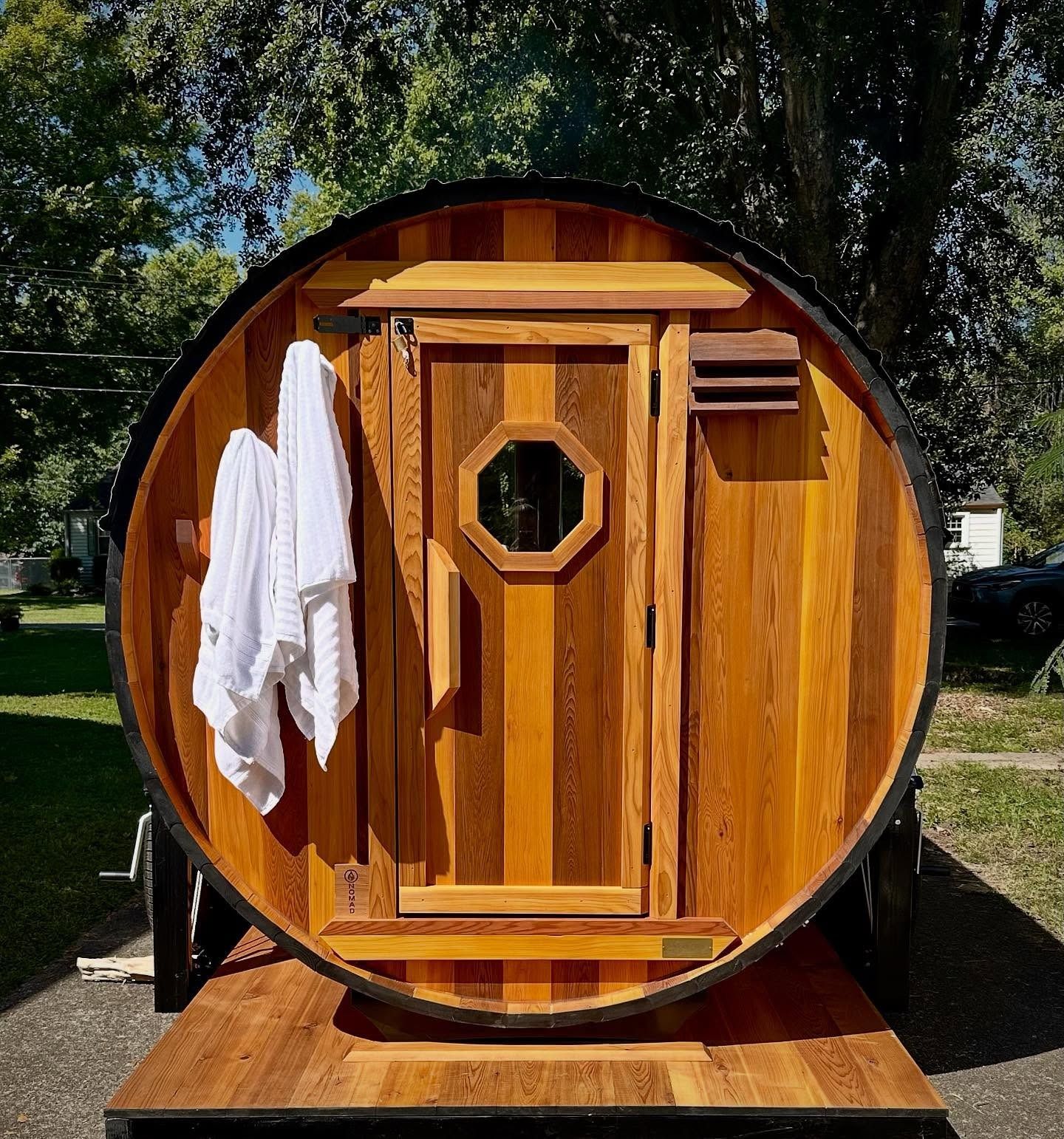 A barrel sauna with wood door, towels, and decorative octagon window. Outdoor setting, parked on a trailer.