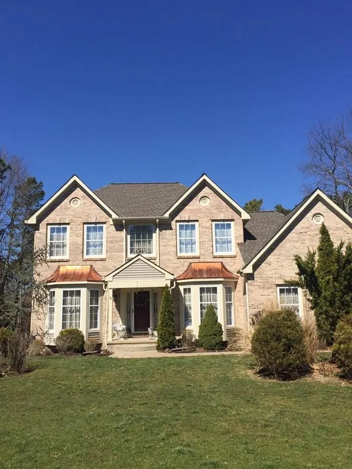 Two-story beige brick house with copper-colored awnings, green lawn, and blue sky.