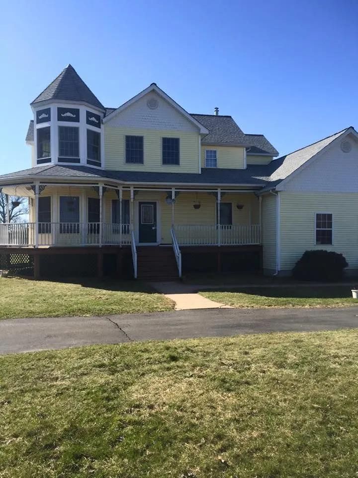 Yellow house with turret and wraparound porch. Driveway in front, blue sky.