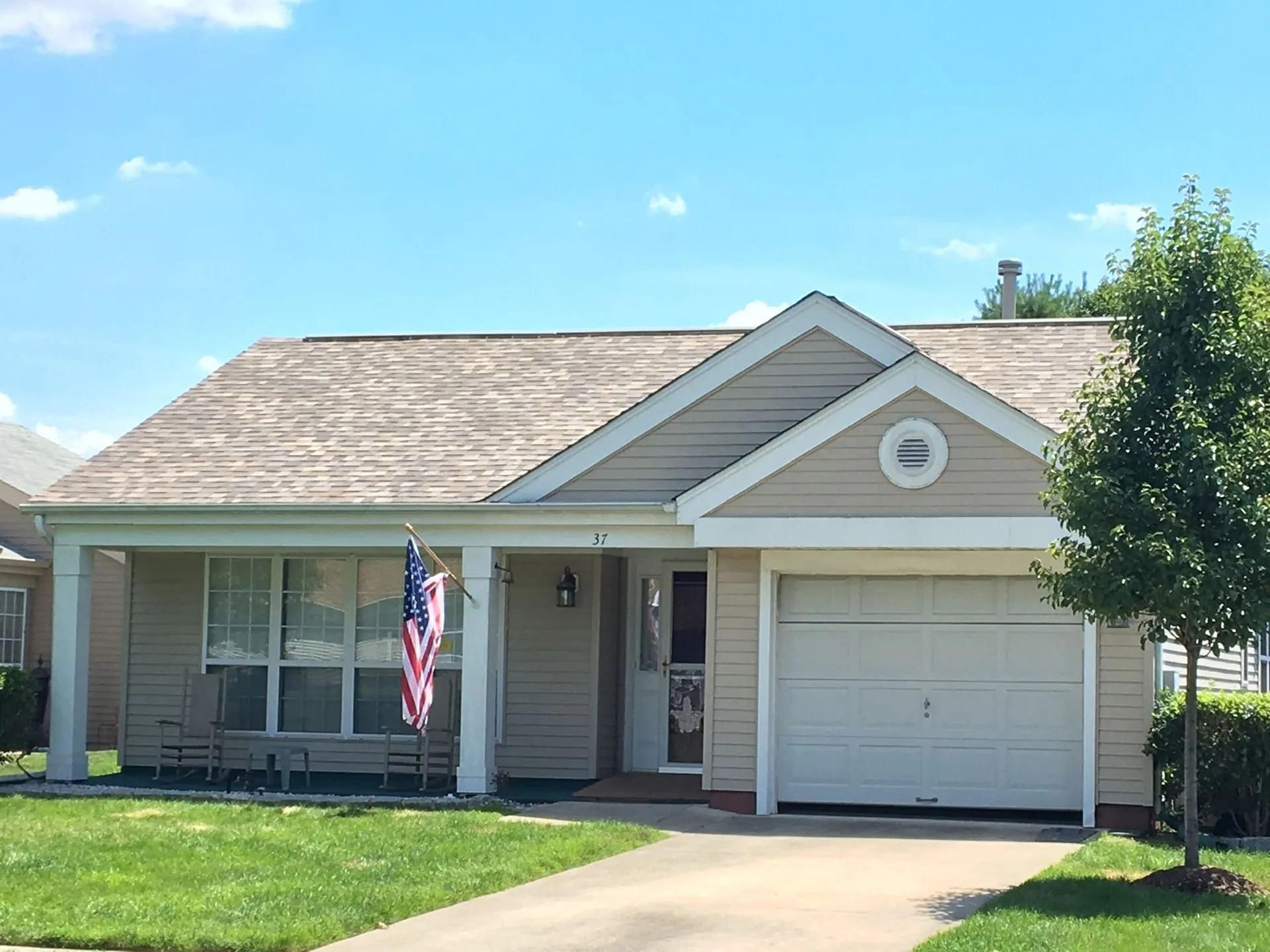 Beige bungalow with a white garage door, porch, and American flag on a sunny day.