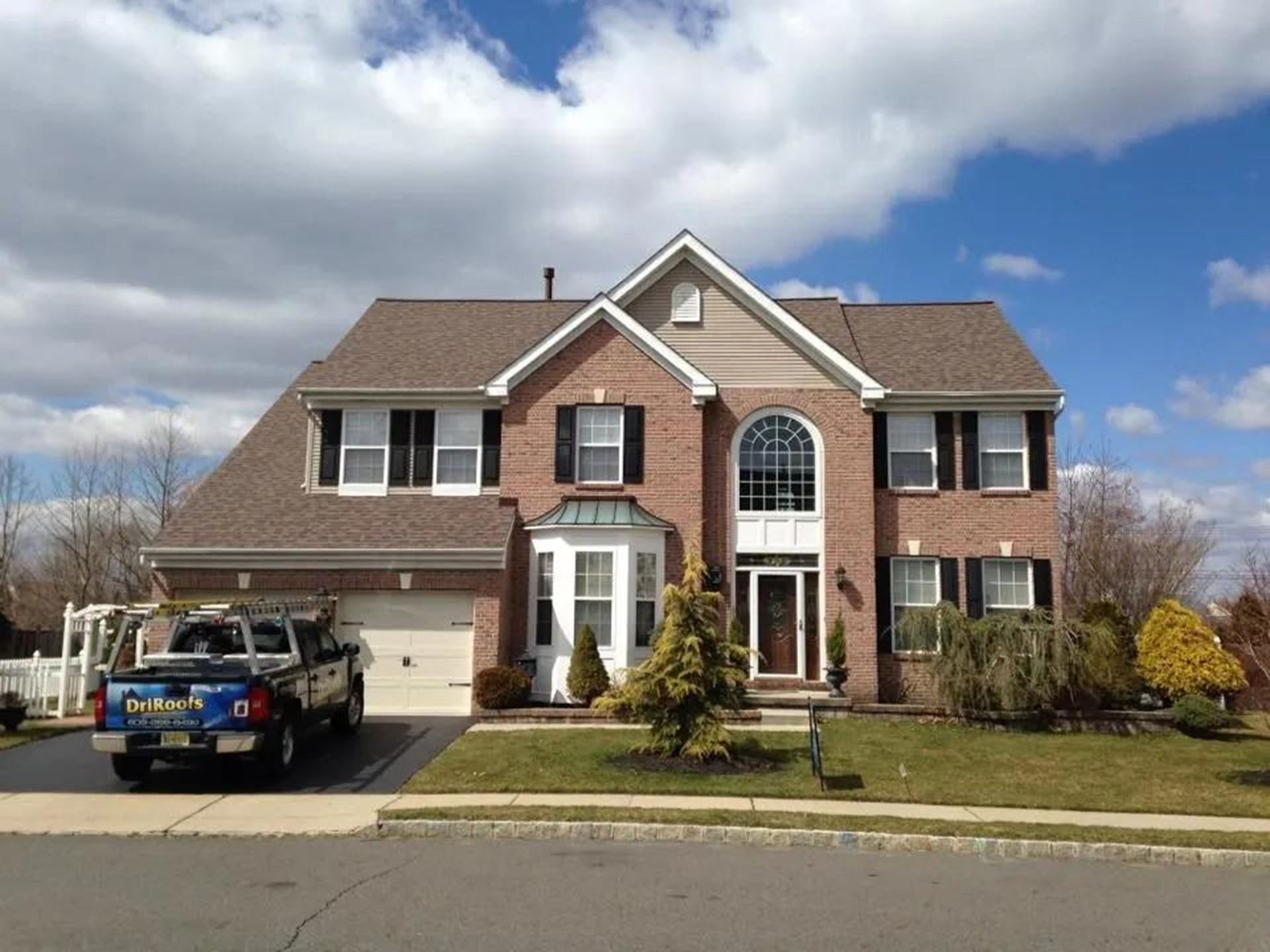 Two-story brick house with brown roof, black shutters, and truck parked in the driveway on a sunny day.