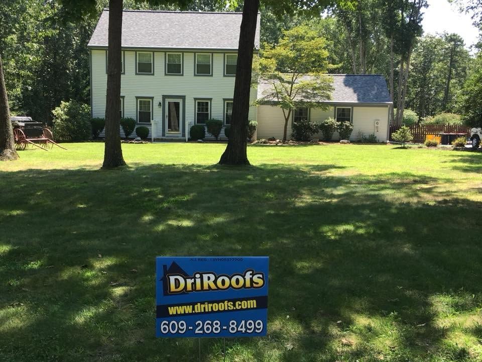 Two-story white house with green shutters in a sunny yard, with a DriRoofs sign in front