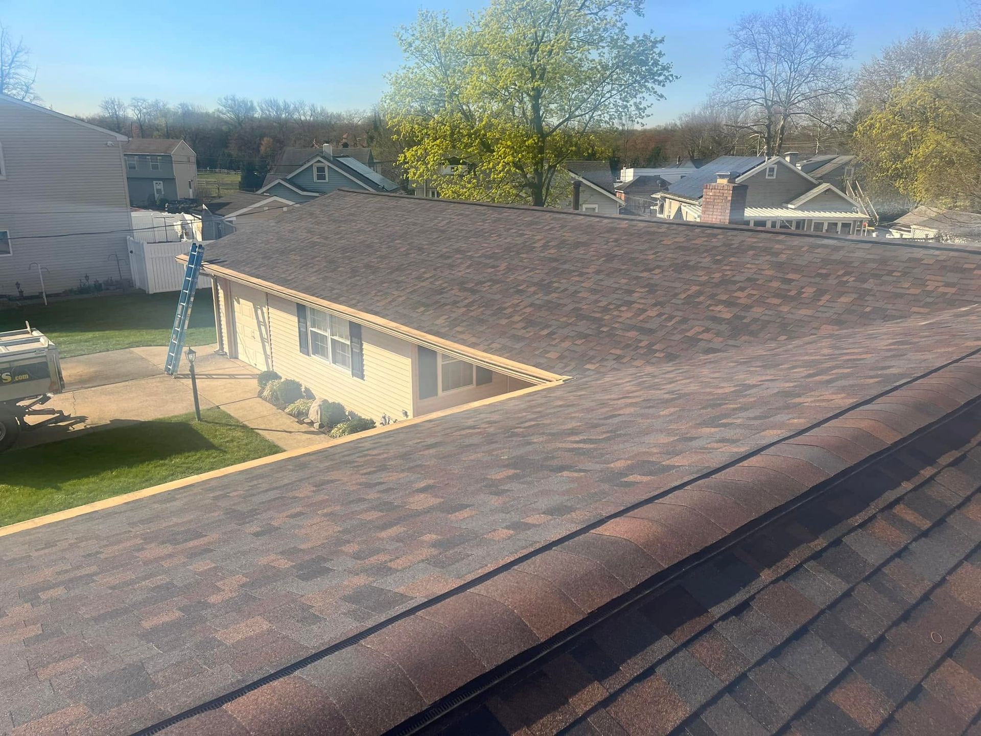 Rooftop view of a brown shingle roof over a suburban backyard with houses and trees in the distance