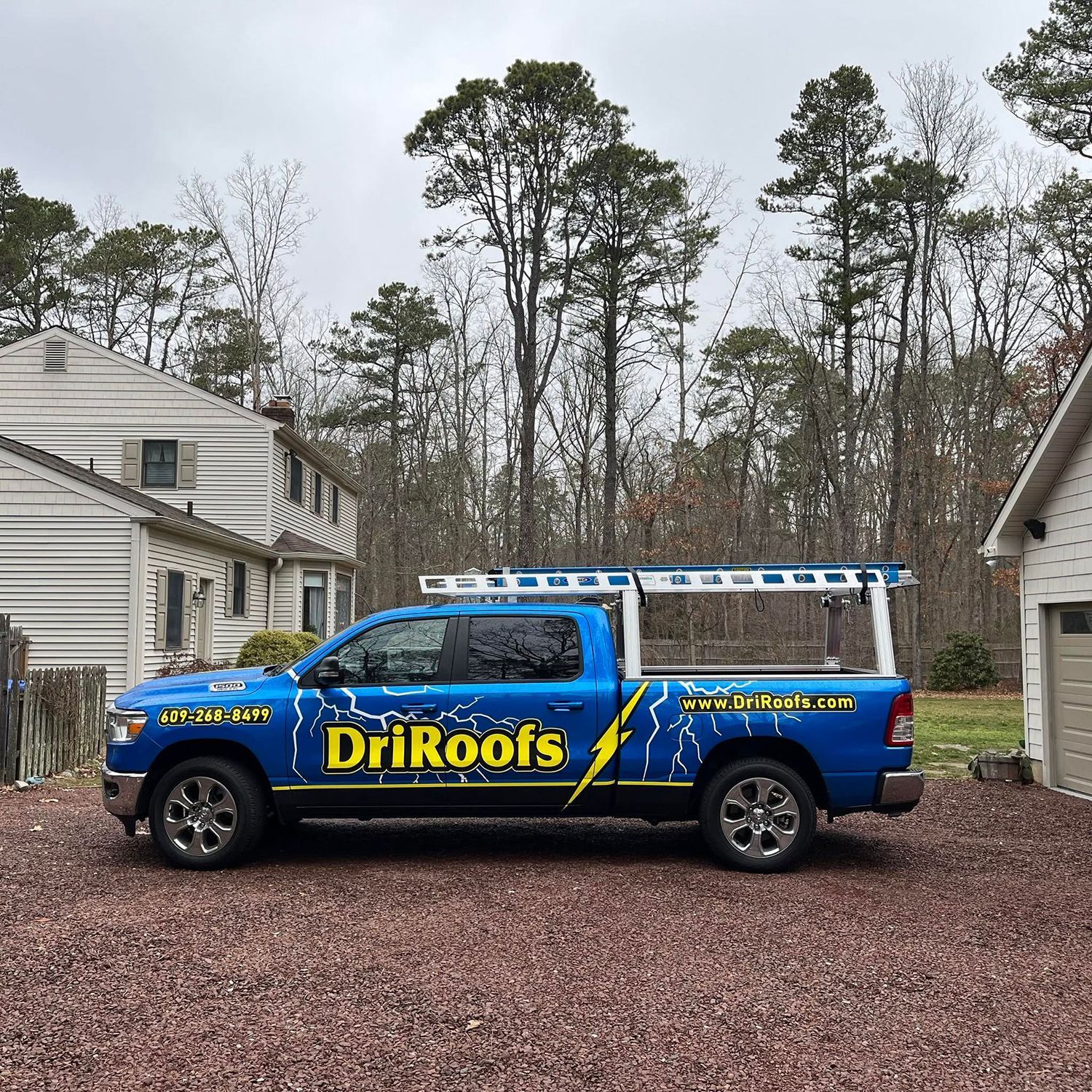 Blue DriRoofs work truck parked in a gravel driveway between houses on an overcast day
