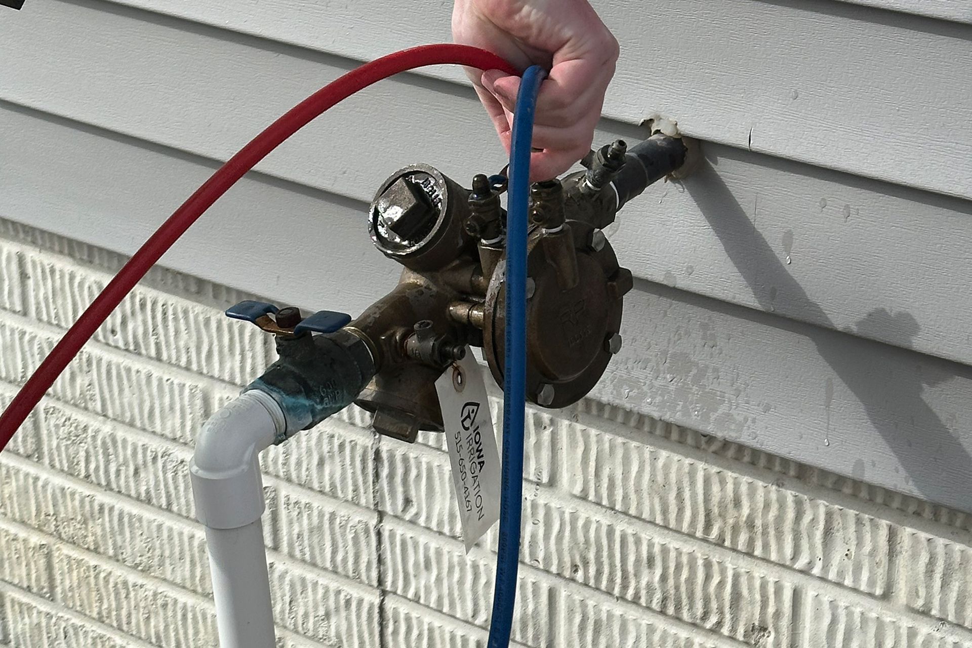 Hand holding blue and red hoses connected to a backflow preventer on a building's exterior.