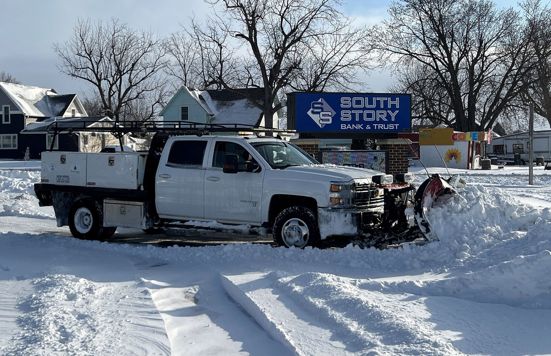 A white truck with a snowplow is clearing snow in front of a building.