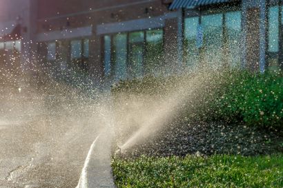 Sprinkler sprays water on green grass, building in background.