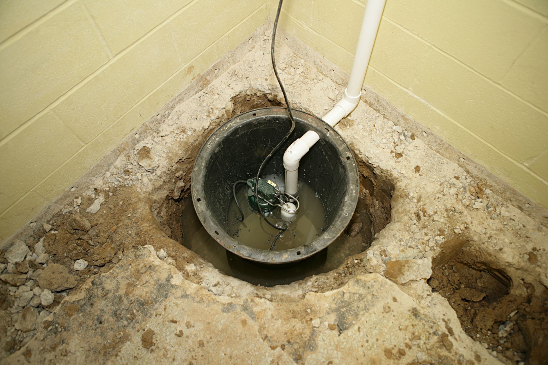 Basement floor with a sump pit, white pipe, and damp, crumbling concrete around it