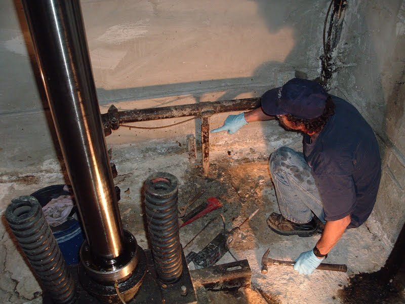 Worker kneeling in a crawlspace beside pipes and rubble, wearing gloves and a cap.