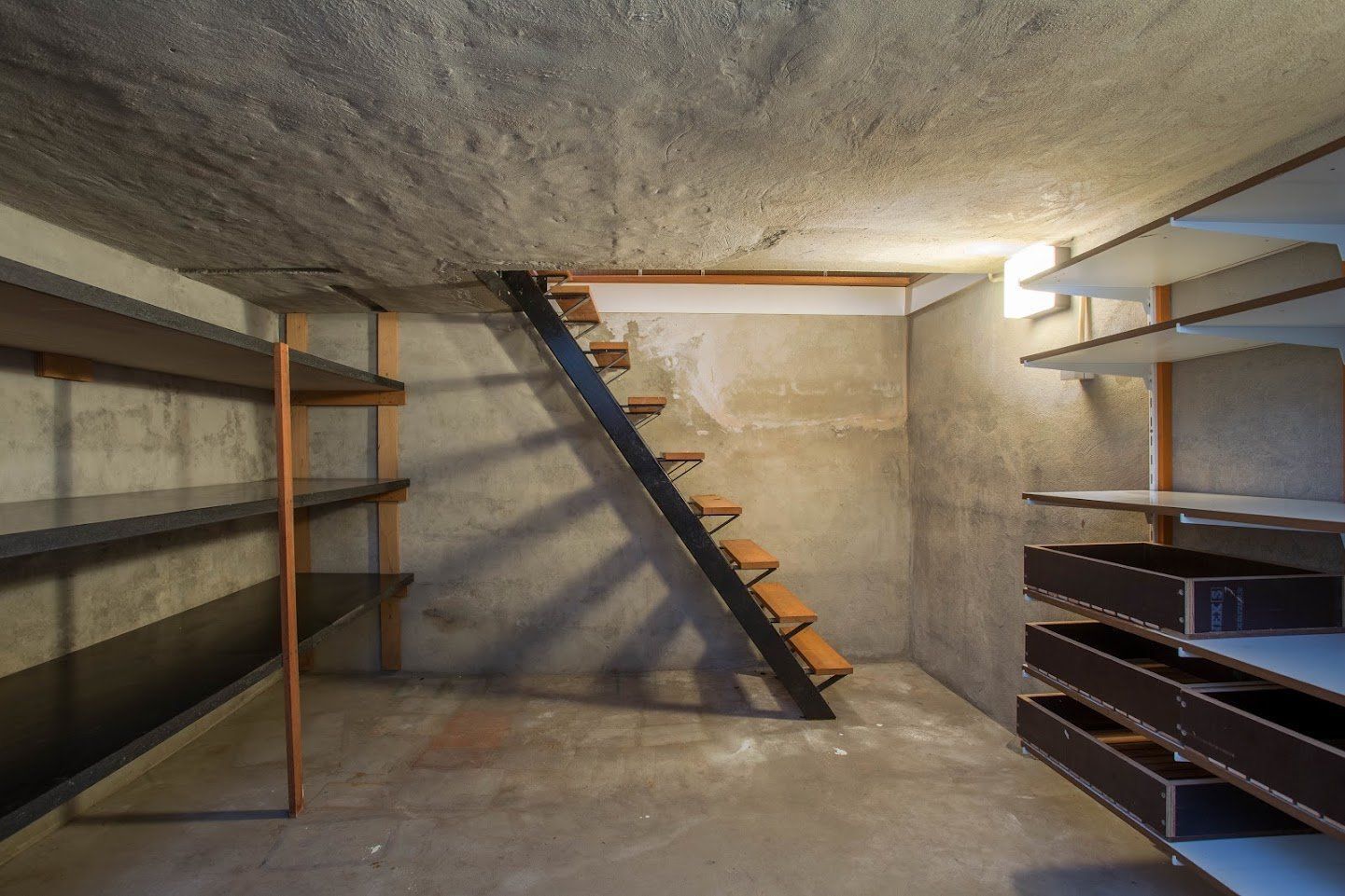 Empty basement with concrete walls, shelves, and a narrow wooden staircase to the upper level