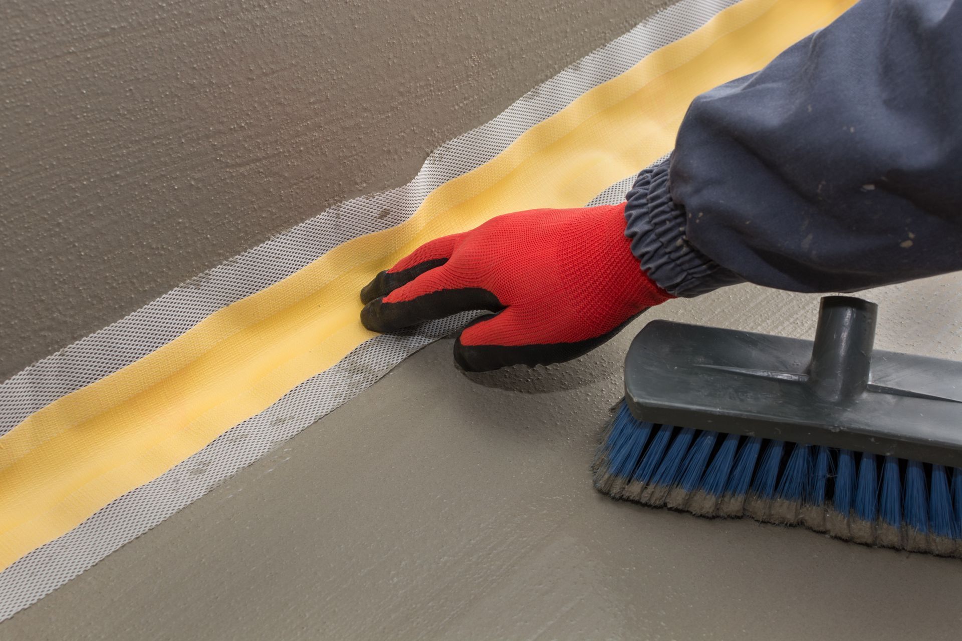 Gloved hand smoothing a gray surface along a yellow seam with a brush nearby