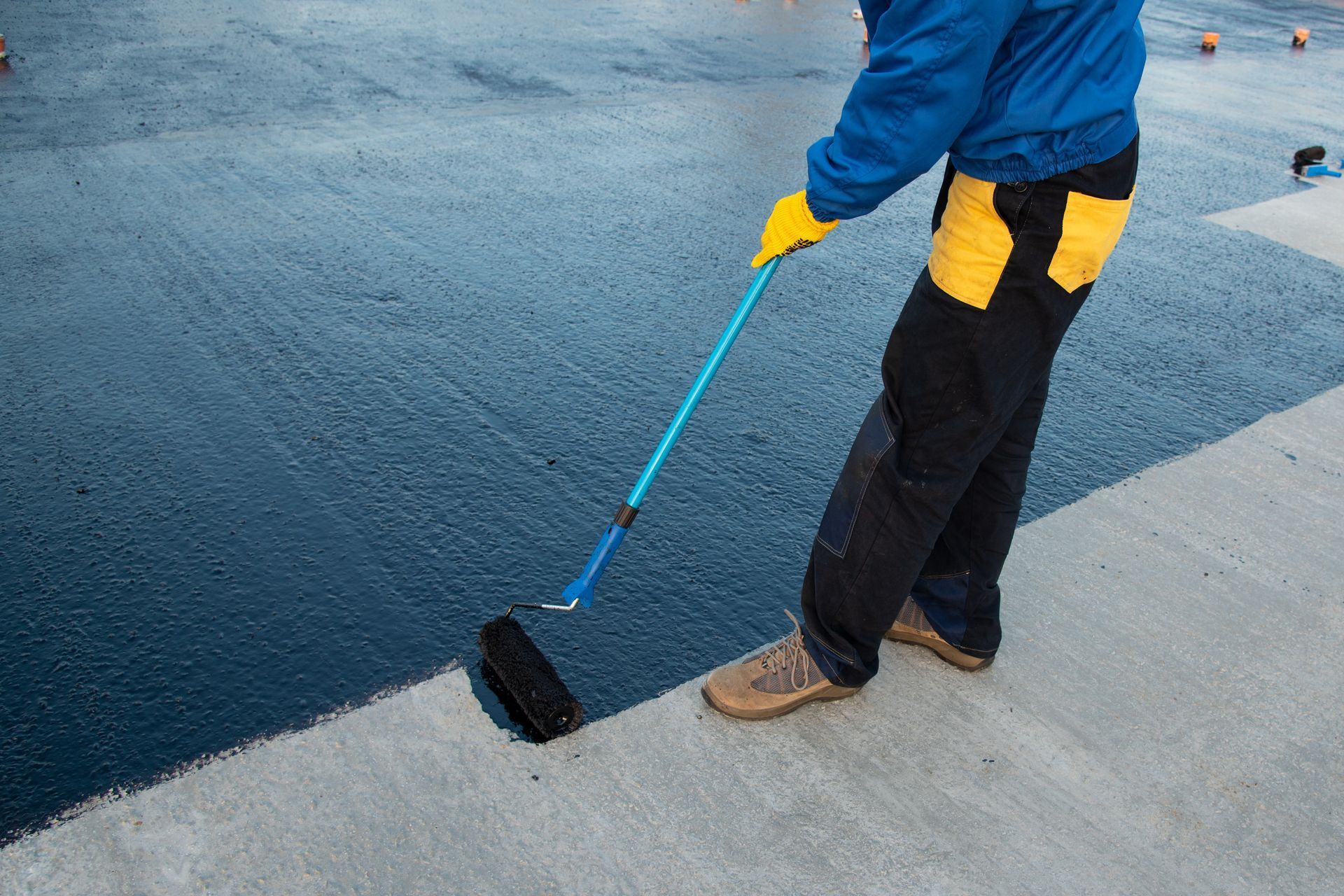 Worker using a roller to paint a dark blue surface beside a light concrete edge