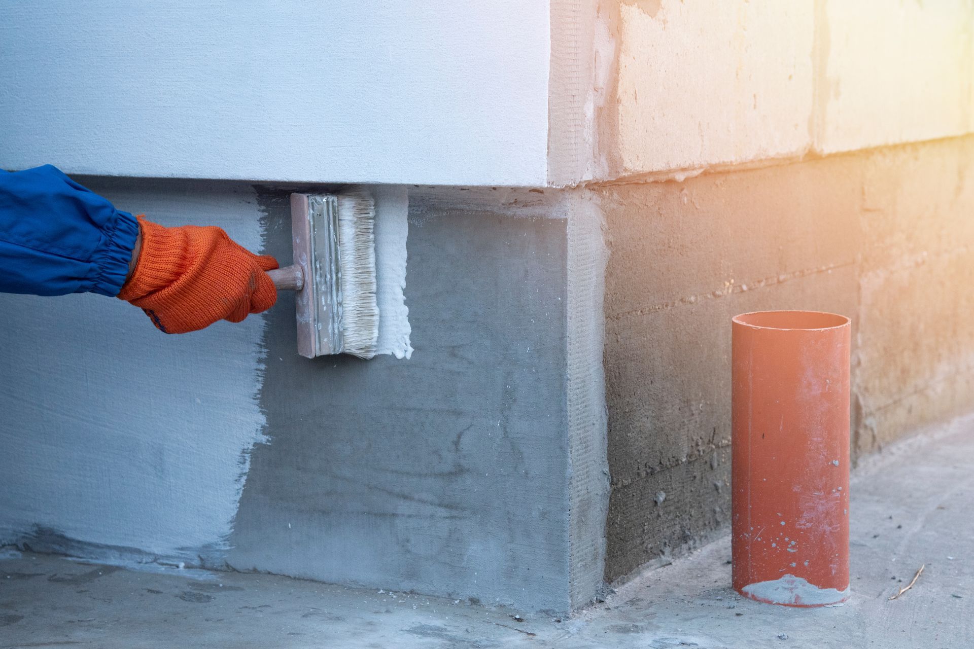 Gloved hand painting a gray wall near a corner with an orange pipe nearby