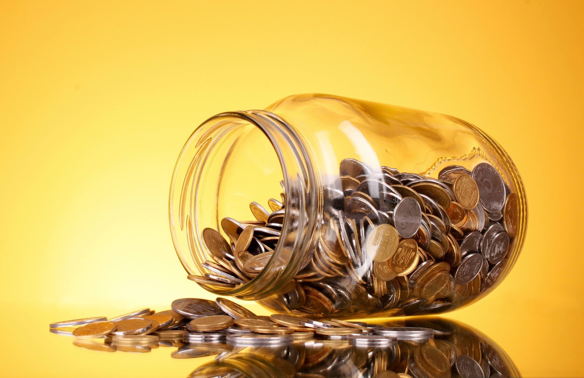 Spilled jar of coins on a yellow background, with coins scattered and reflected on the surface
