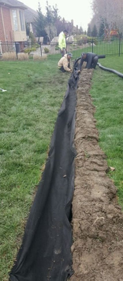 Black geotextile lining a long trench in a grassy yard, with workers in the distance.