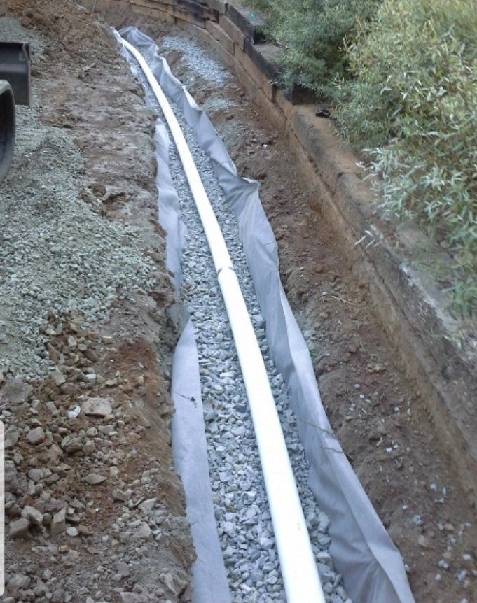 A white pipe laid in a narrow trench lined with gray fabric, beside dirt and vegetation.