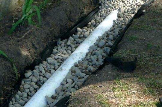 White drainage pipe in a rocky trench beside grass and soil.