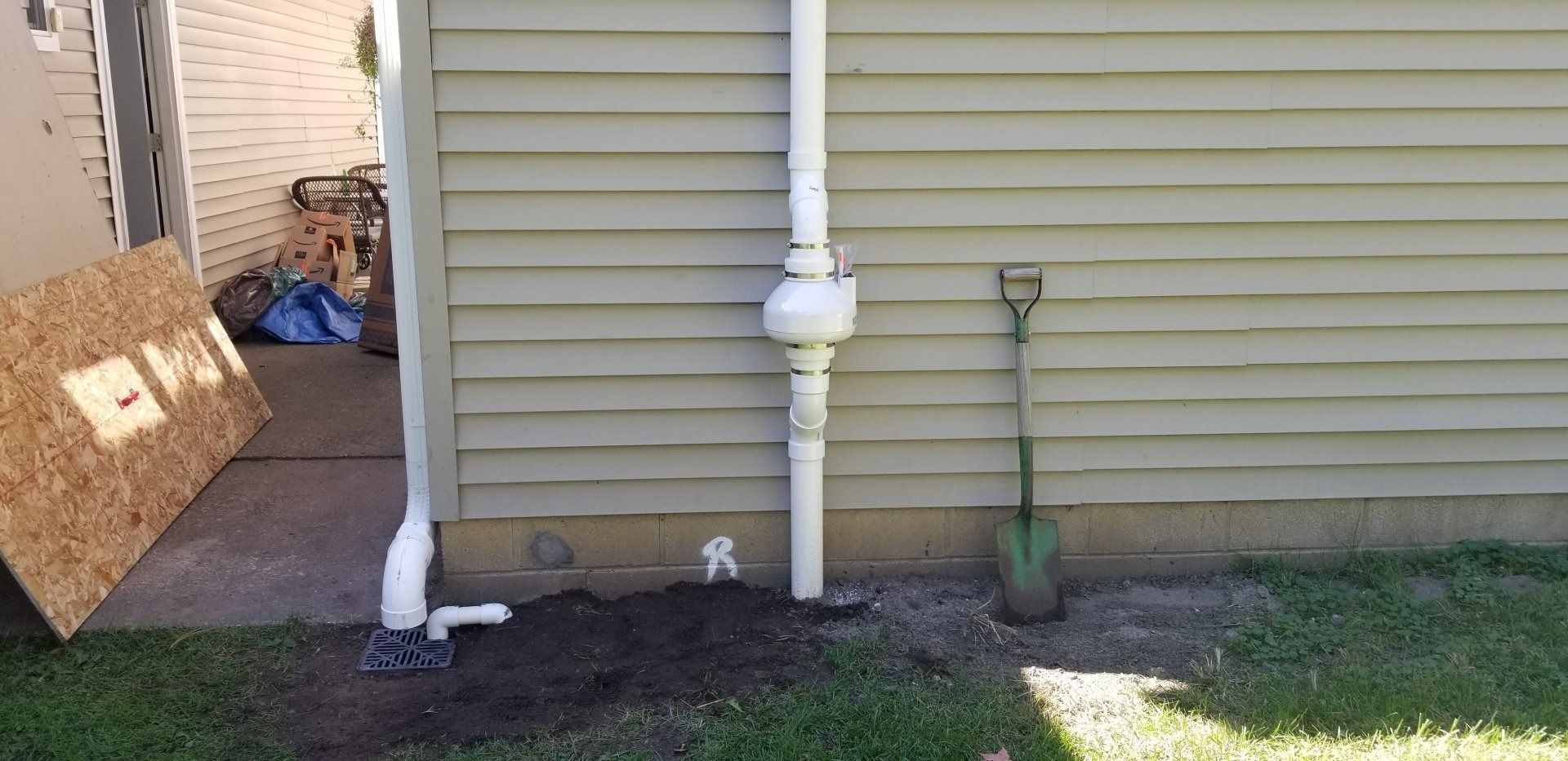 House siding with white pipes, utility meter, and small shovel beside a concrete foundation.