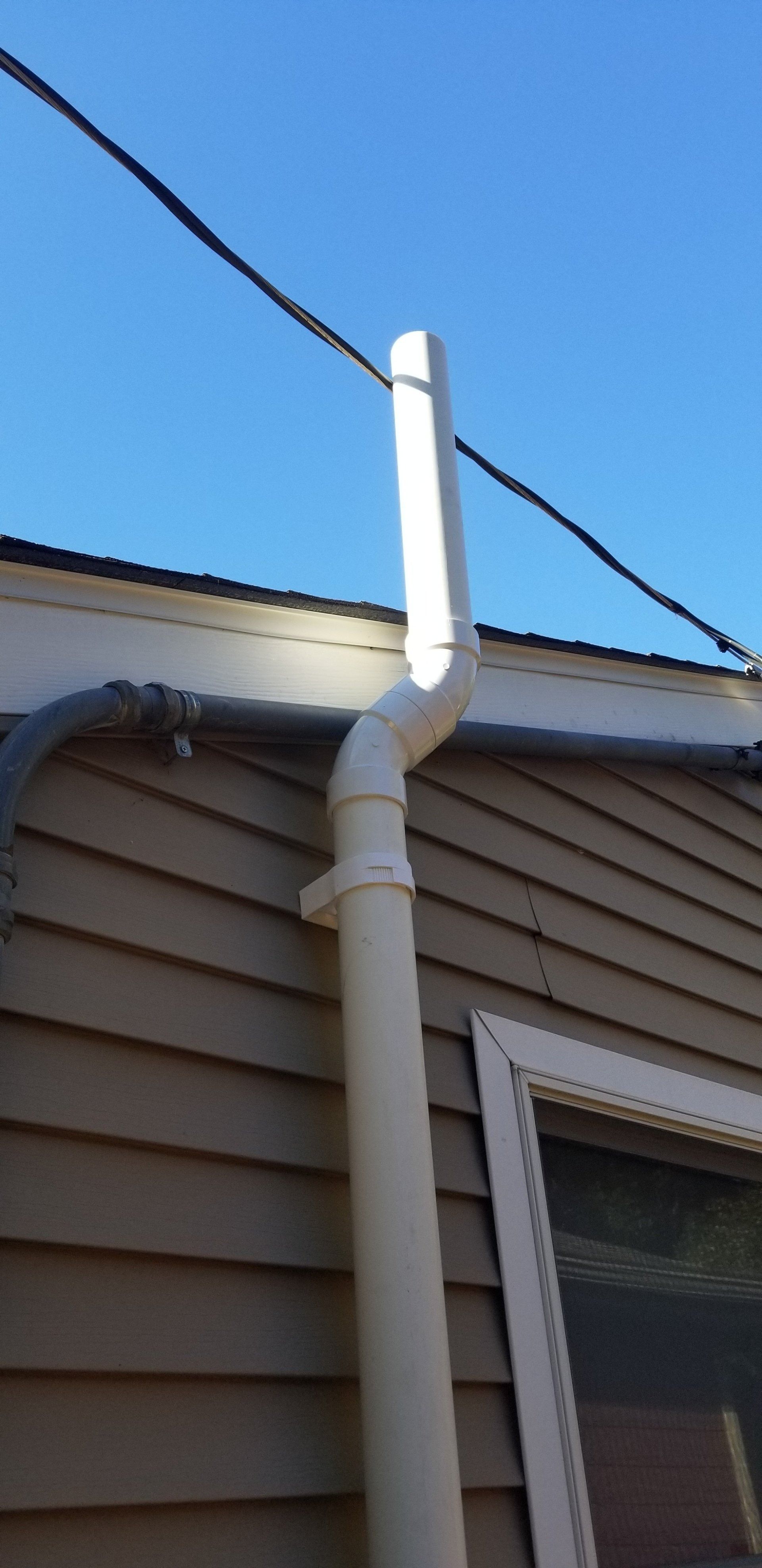 White downspout and elbow pipe on the side of a beige house beneath a black cable