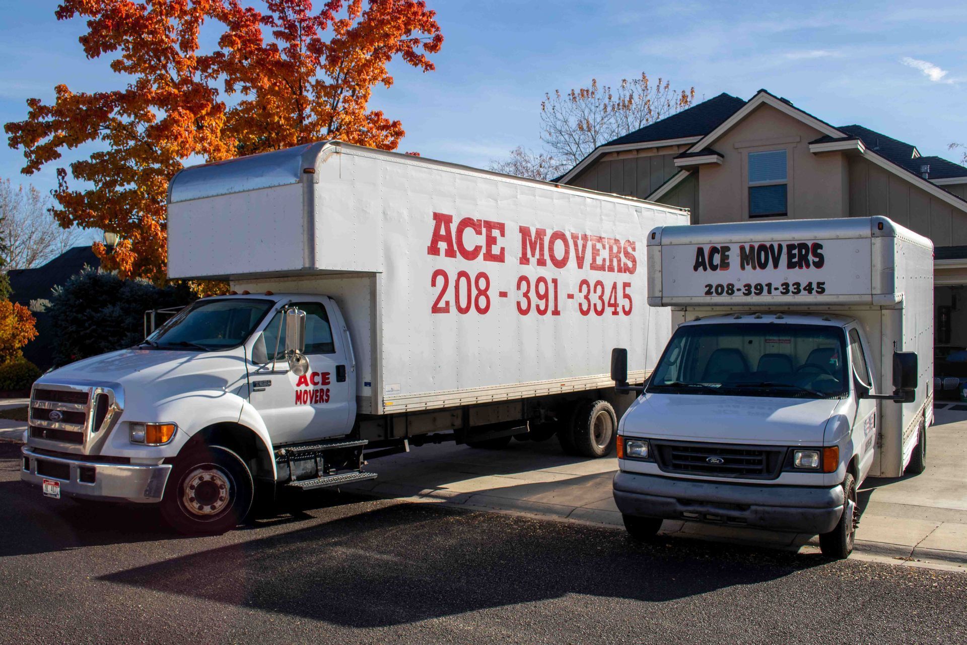 Two white moving trucks parked in front of a house, with autumn foliage in the background.
