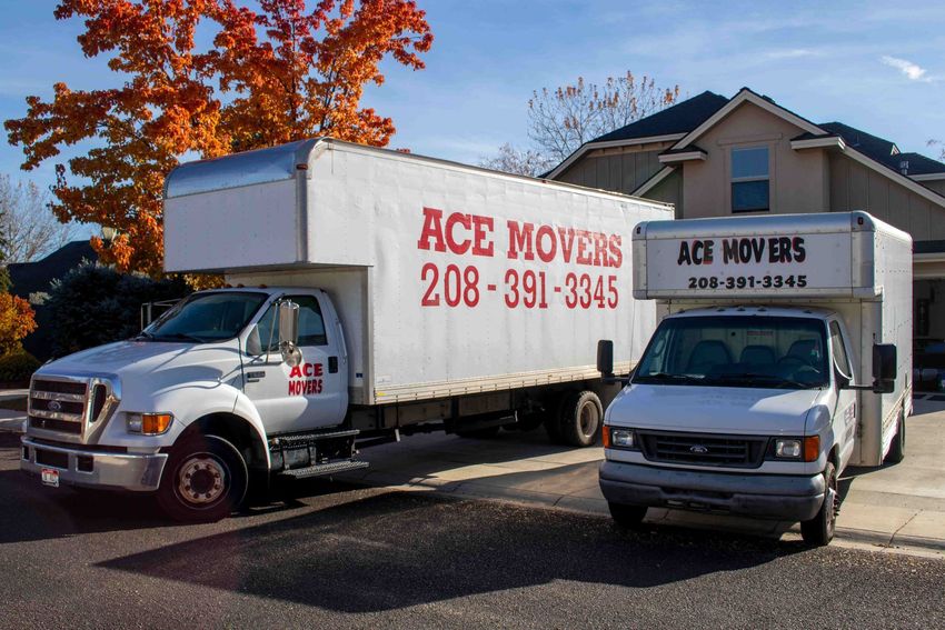 Two white moving trucks parked in front of a house, with autumn foliage in the background.