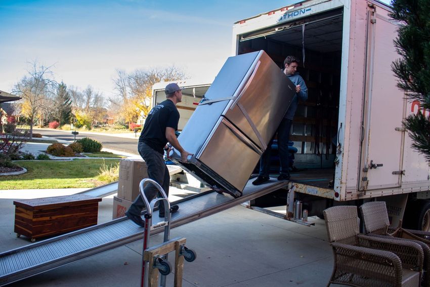 Two movers load a stainless steel refrigerator into a moving truck on a sunny day.