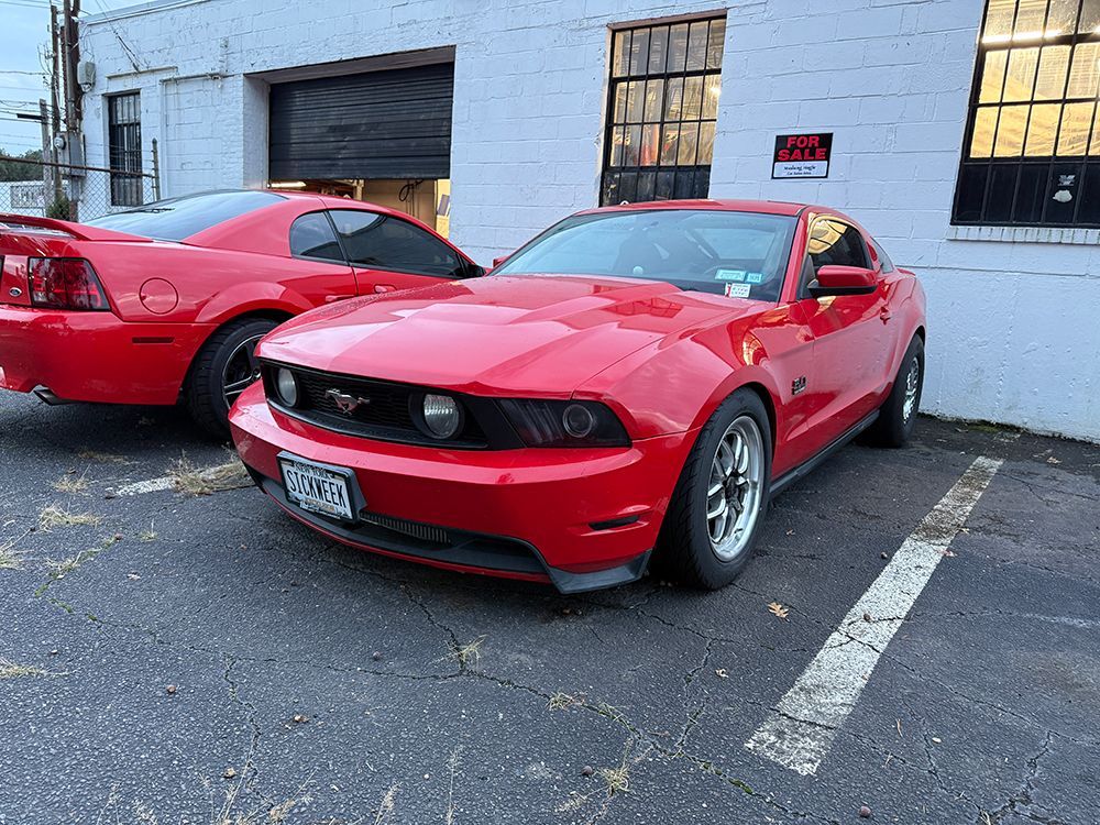 Red Ford Mustang in front of a white building, another red Mustang behind it.