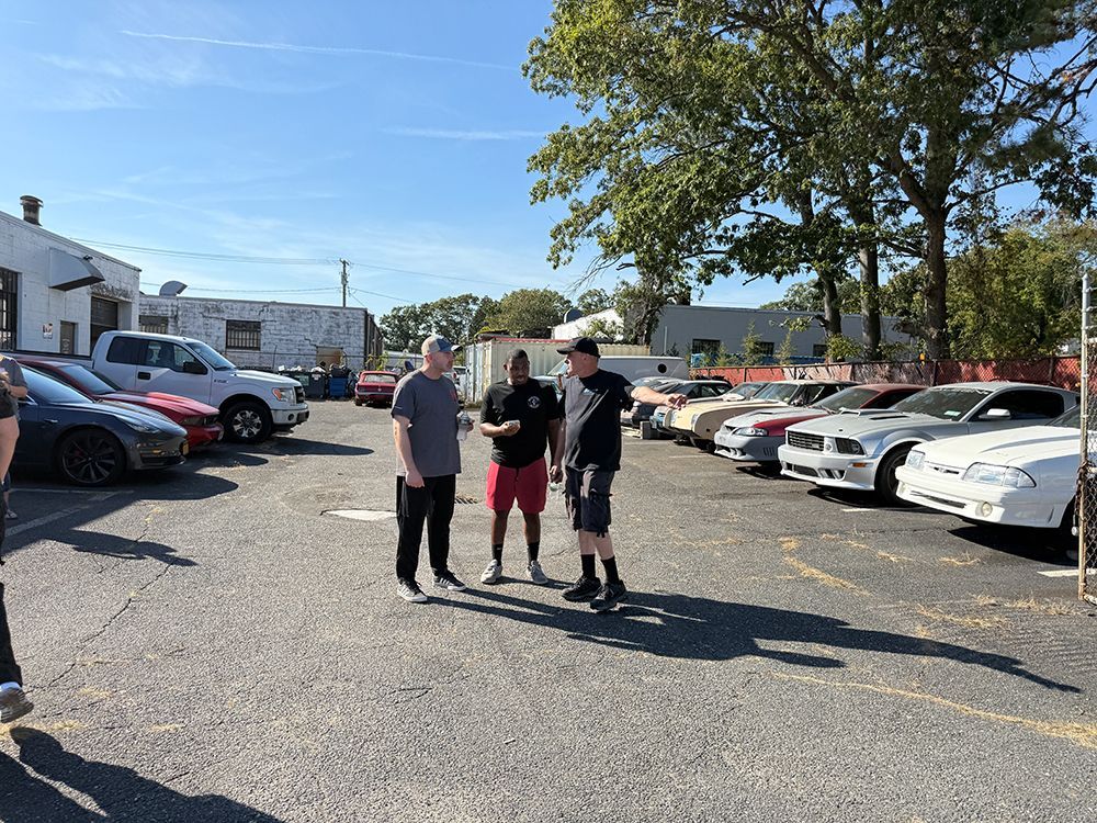 Three people talking in a parking lot with various cars parked around them on a sunny day.