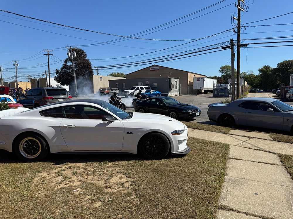 White Mustang and other cars at a car event, with smoke rising in the air, outdoors on a sunny day.