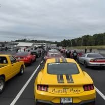 Yellow Mustang race car in a lineup with other vehicles at a racetrack.